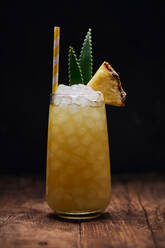 Wooden table with glass of yellow cocktail with ice cubes and refreshing cocktail garnished with spiky leaves and striped straw - ADSF24972