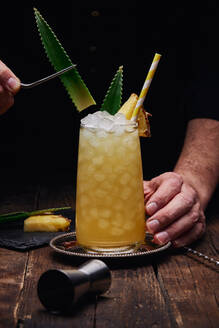 Crop anonymous barman garnishing alcohol cocktail with green leaves and pineapple piece on tray near shot glass at wooden table on black background - ADSF24973