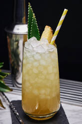 Yellow cocktail in glass garnished with pineapple piece and green leaves with paper straw placed on slate coaster with bar spoon - ADSF24975
