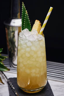 Gelber Cocktail im Glas, garniert mit Ananasstück und grünen Blättern, mit Papierstrohhalm auf Schieferuntersetzer mit Barlöffel - ADSF24975