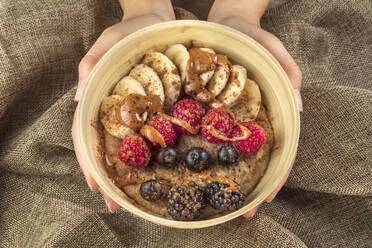 Top view of crop anonymous cook showing bowl with banana slices and assorted fresh berries with caramel sauce for breakfast - ADSF24994