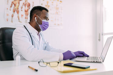 Konzentrierter afroamerikanischer Arzt in medizinischer Kutte und Maske, der in einer modernen Klinik an einem Laptop arbeitet - ADSF25008