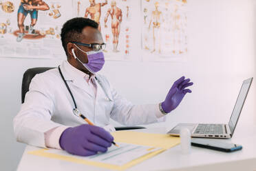 African American doctor in eyeglasses working with online patient on netbook while writing in patient file at table in hospital - ADSF25009