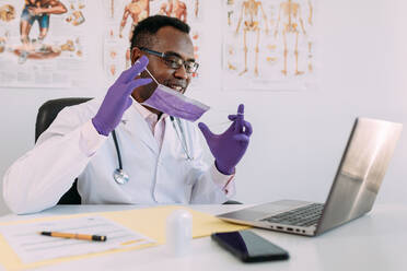 Ein afroamerikanischer Arzt mit Brille und Gesichtsmaske arbeitet mit einem Online-Patienten an einem Netbook, während er in einer Patientenakte an einem Tisch im Krankenhaus schreibt - ADSF25010