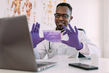 Positiv erwachsener afroamerikanischer männlicher Arzt in medizinischer Uniform und Latexhandschuhen, der lächelt und eine Maske zeigt, während er eine Videokonferenz über einen Laptop in einem modernen Krankenhaus abhält - ADSF25012