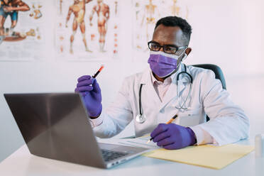 Nicht erkennbarer ernsthafter schwarzer Wissenschaftler in medizinischer Robe und Maske, der eine Blutprobe in einem Reagenzglas demonstriert, während er einen Videogespräch auf einem Laptop in einer Klinik führt - ADSF25015