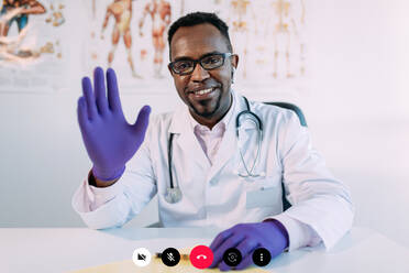 Fröhlicher afroamerikanischer Arzt in medizinischer Uniform und Brille, der lächelt und mit der Hand in Richtung Kamera winkt, während er in einem modernen Labor während einer Videokonferenz am Tisch sitzt - ADSF25016