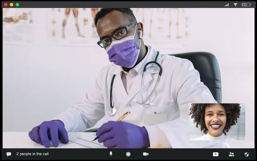 Concentrated African American male doctor in medical robe and mask looking at camera while communicating with positive female during video call - ADSF25025