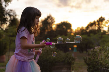 Girl playing with soap bubbles while holding wand during sunset - GEMF04876