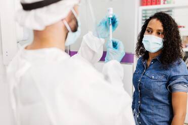 Doctor in latex gloves and face shield filling in syringe from bottle with vaccine preparing to vaccinate unrecognizable female African American patient in clinic during coronavirus outbreak - ADSF25027