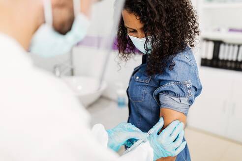 Cropped unrecognizable male medical specialist in protective uniform, latex gloves and face shield vaccinating African American female patient in clinic during coronavirus outbreak - ADSF25029