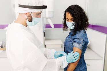 Side view of male medical specialist in protective uniform, latex gloves and face shield vaccinating African American female patient in clinic during coronavirus outbreak - ADSF25030