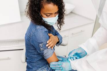 Cropped unrecognizable male medical specialist in protective uniform and latex gloves vaccinating African American female patient in clinic during coronavirus outbreak - ADSF25034