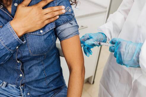 Cropped unrecognizable male medical specialist in protective uniform and latex gloves vaccinating African American female patient in clinic during coronavirus outbreak - ADSF25038