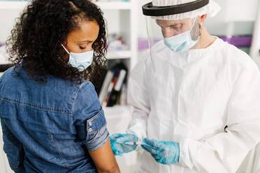 Side view of male medical specialist in protective uniform, latex gloves and face shield vaccinating African American female patient in clinic during coronavirus outbreak - ADSF25041