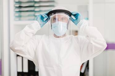 Positiver männlicher Arzt mit Latexhandschuhen und weißer Uniform, der eine Gesichtsschutzmaske aufsetzt, in einem modernen medizinischen Büro steht und in die Kamera schaut - ADSF25044