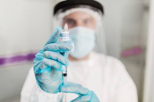 Blurred unrecognizable medic in protective face shield mask and latex gloves with vial of coronavirus vaccine and syringe showing to camera while standing in hospital room - ADSF25046