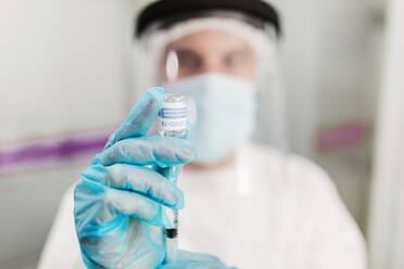 Unscharfer, nicht erkennbarer Sanitäter mit Gesichtsschutzmaske und Latexhandschuhen, der eine Ampulle mit Coronavirus-Impfstoff und eine Spritze in die Kamera hält, während er in einem Krankenhauszimmer steht - ADSF25047