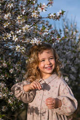 Adorable lächelndes kleines Kind in Kleid stehend in der Nähe blühenden Baum mit Blumen im Frühling Park und schauen weg - ADSF25055