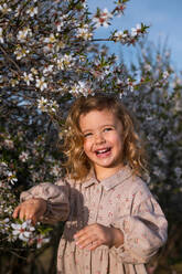 Adorable lächelndes kleines Kind in Kleid stehend in der Nähe blühenden Baum mit Blumen im Frühling Park und schauen weg - ADSF25056