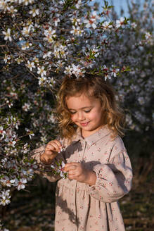 Adorable lächelndes kleines Kind im Kleid stehend in der Nähe blühenden Baum mit Blumen im Frühling Park und Blick nach unten - ADSF25057