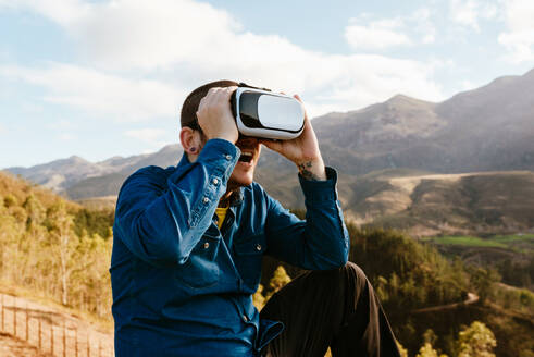 Neugieriger männlicher Reisender, der auf einem Hügel sitzt und die virtuelle Realität mit einer Brille in den Bergen an einem sonnigen Tag erlebt - ADSF25066