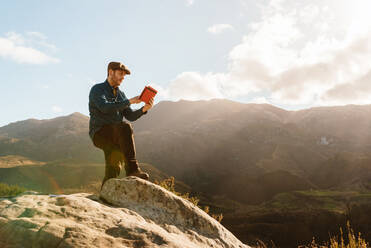 Ruhiger männlicher Entdecker mit Tablet, der auf einem Felsen steht und die spektakuläre Aussicht auf die Berge an einem sonnigen Tag genießt - ADSF25069