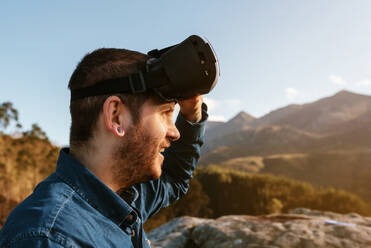 Curious male traveler sitting on hill and experiencing virtual reality in goggles in mountains on sunny day - ADSF25070