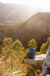 Ruhiger männlicher Forscher mit Tablet auf einem Hügel in bergigem Gelände bei Sonnenuntergang sitzend - ADSF25072