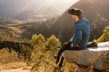 Side view of male traveler in VR glasses interacting with virtual reality while sitting on hill in mountainous terrain at sunset - ADSF25073
