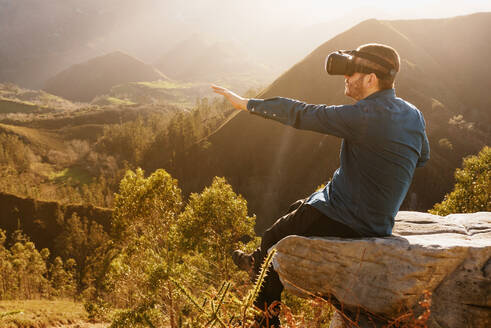 Side view of male traveler in VR glasses interacting with virtual reality while sitting on hill in mountainous terrain at sunset - ADSF25074