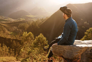 Side view of male traveler in VR glasses interacting with virtual reality while sitting on hill in mountainous terrain at sunset - ADSF25075