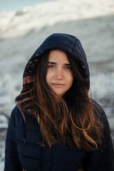 Junge sanfte weibliche Reisende mit Kapuze schaut in die Kamera gegen verschneite Berge im Winter an einem sonnigen Tag - ADSF25077