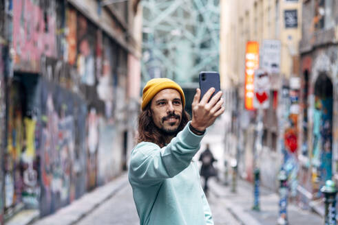 Zufriedener Mann mit Bart in Freizeitkleidung, der ein Selbstporträt mit dem Handy in der Stadt vor einem unscharfen Hintergrund aufnimmt - ADSF25082