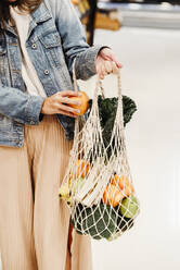 Cropped unrecognizable female standing with assorted fruits and vegetables in eco friendly mesh bag against blue wall in city - ADSF25089