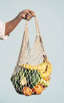 Cropped unrecognizable female standing with assorted fruits and vegetables in eco friendly mesh bag against blue wall in city - ADSF25091