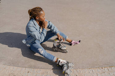 Eine afroamerikanische Frau in Jeanskleidung zieht an einem sonnigen Tag im Skatepark ihre Rollerblades an - ADSF25098