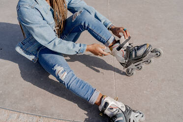 Eine afroamerikanische Frau in Jeanskleidung zieht an einem sonnigen Tag im Skatepark ihre Rollerblades an - ADSF25100