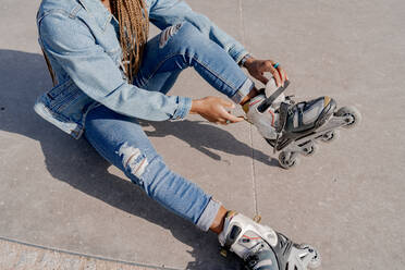 From above of African American female in denim outfit putting on rollerblades on sunny day in skate park - ADSF25100
