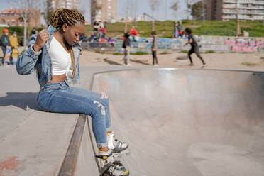 Coole schwarze Frau mit geflochtener Frisur und in Rollerblades sitzt auf der Rampe im Skatepark und schaut nach unten - ADSF25101