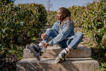 Smiling African American female putting on rollerblades while sitting on stairs in summer park and looking away - ADSF25105