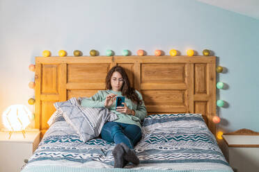 Junge Frau mit türkisfarbenem Sweatshirt und Brille liegt auf dem Bett und benutzt das Mobiltelefon - ADSF25137
