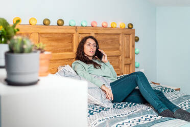 Junge Frau mit türkisfarbenem Sweatshirt und Brille liegt auf dem Bett und telefoniert mit dem Handy - ADSF25138