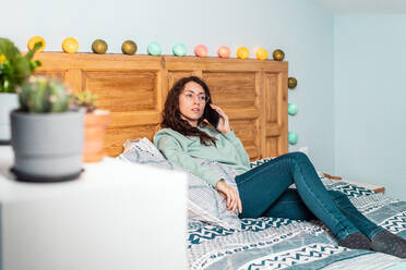 Young Woman with a turquoise sweatshirt and glasses lying on bed talking on the mobile phone - ADSF25138