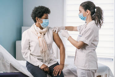 Medizinische Fachkraft in Schutzkleidung, Latexhandschuhen und Gesichtsmaske bei der Impfung einer reifen afroamerikanischen Patientin in einer Klinik während des Ausbruchs des Coronavirus - ADSF25145