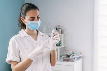 Female doctor in latex gloves and face shield filling in syringe from bottle with vaccine preparing to vaccinate patient in clinic during coronavirus outbreak - ADSF25150