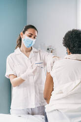 Medizinische Fachkraft in Schutzkleidung, Latexhandschuhen und Gesichtsmaske bei der Impfung einer reifen afroamerikanischen Patientin in einer Klinik während des Ausbruchs des Coronavirus - ADSF25154