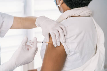 Abgeschnittene, nicht erkennbare weibliche medizinische Fachkraft in Schutzkleidung, Latexhandschuhen und Gesichtsmaske bei der Impfung einer anonymen afroamerikanischen reifen Patientin in einer Klinik während des Ausbruchs des Coronavirus - ADSF25155