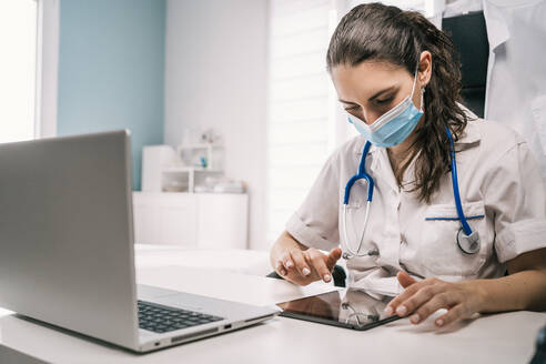 Konzentrierte junge Ärztin in medizinischer Maske und Uniform mit Stethoskop, die an einem Tisch sitzt und auf einem Tablet surft, während sie in einem modernen Arbeitsraum arbeitet - ADSF25157