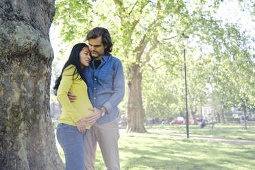 Frau umarmt ihren Mann vor einem Baumstamm im Park - ASGF00601